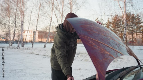 Young man in hooded winter jacket opens car bonnet and places support rod to secure hood in snowy outdoor area surrounded by leafless trees and distant buildings photo