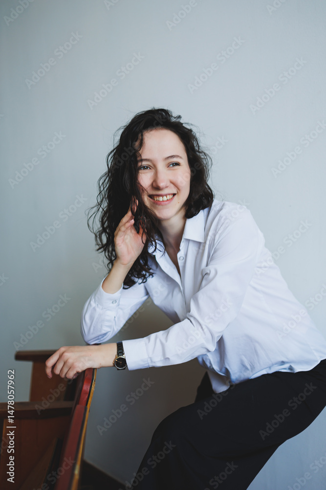 Confident Young Woman in Professional Attire Laughing while Posing with a Wooden Chair in a Minimalist Setting