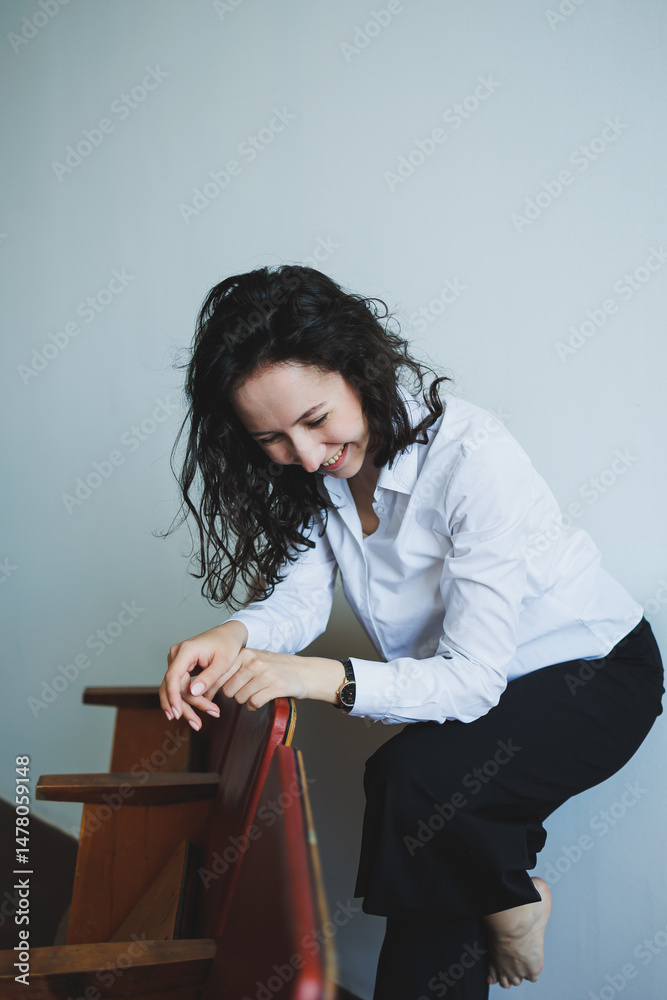 A cheerful woman in an elegant white shirt playfully interacts with trendy furniture in her modern, minimalist living space, showcasing her vibrant happiness and creative lifestyle