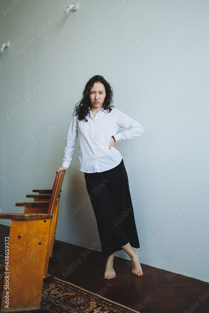 An Elegant Woman in Casual Style is Posing Gracefully in a Minimalist Interior Setting Against a Soft Light Wall, Showcasing a Unique Blend of Comfort and Fashion in Contemporary Design