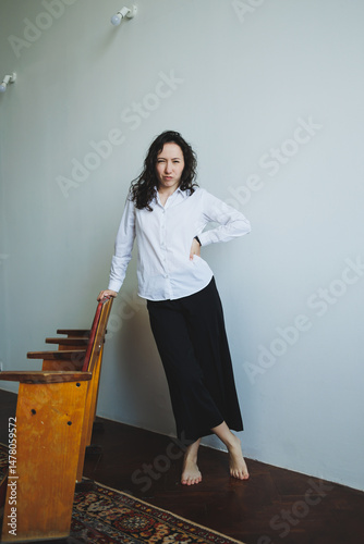 Obraz na płótnie An Elegant Woman in Casual Style is Posing Gracefully in a Minimalist Interior Setting Against a Soft Light Wall, Showcasing a Unique Blend of Comfort and Fashion in Contemporary Design w salonie