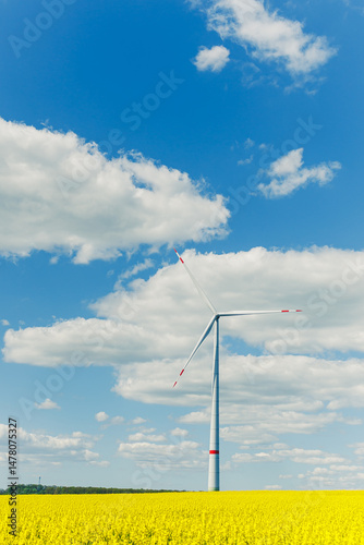 Obraz na płótnie A tall wind turbine stands against a blue sky, surrounded by yellow flowers, symbolizing sustainability and harmony with nature, showcasing commitment to ecological balance and renewable energy w salonie