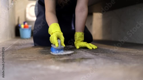 A person kneels on the floor, using a brush to scrub the surface while wearing yellow rubber gloves. The area appears tidy, suggesting a cleaning activity in progress.