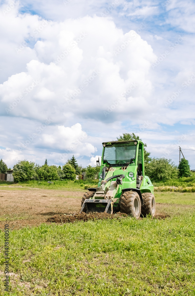 Naklejka premium Mini skid steer loader clear a construction site. Small tractor with a ground leveler moves soil. Land work by the territory improvement, plot leveling. Plowing agricultural field. High quality photo.