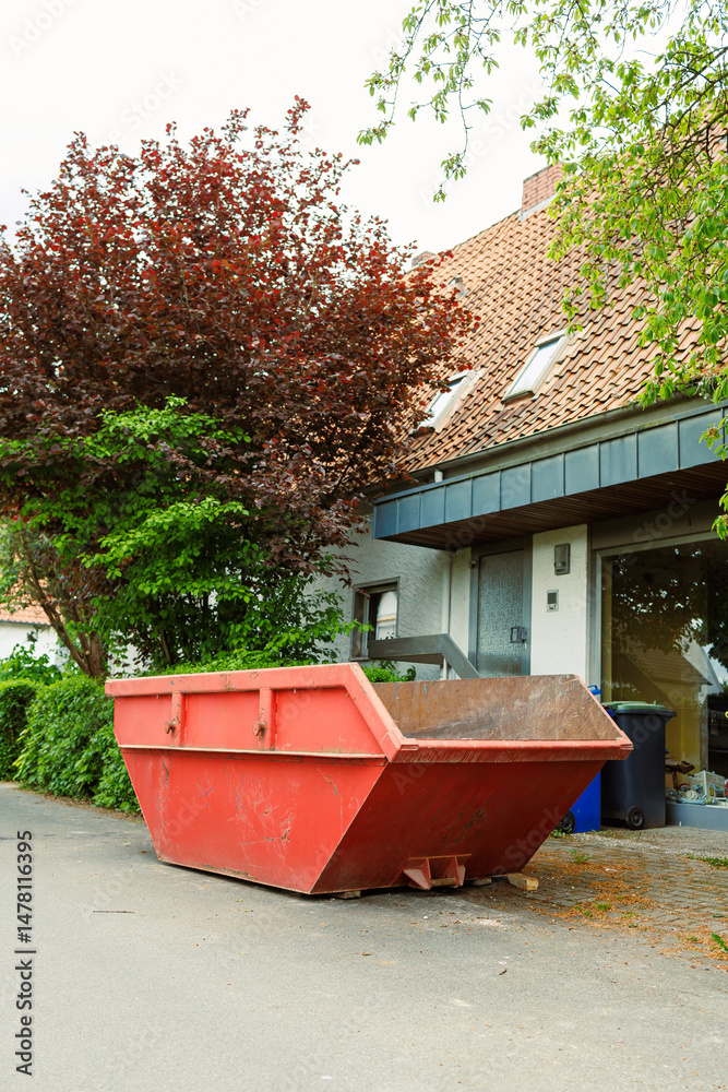 A vibrant red construction dumpster vividly stands out on a serene and quiet street, surrounded by lush greenery and a contemporary home that enhances the neighborhoods peaceful atmosphere and charm