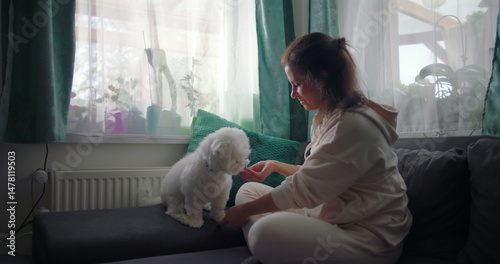 Woman sitting on couch petting white dog in cozy home setting, soft cushions and bright windows enhancing a warm and calm indoor atmosphere
