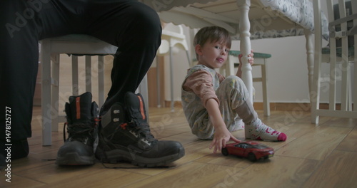 Young boy sitting on the floor with a toy car, next to sturdy boots and a dining chair, in a cozy home with wooden flooring and a warm atmosphere