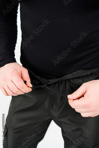 Obraz na płótnie A fashionable male model is seen adjusting the waistband of his sleek black athletic pants, all set against a clean white background that enhances the focus on his outfit and style w salonie