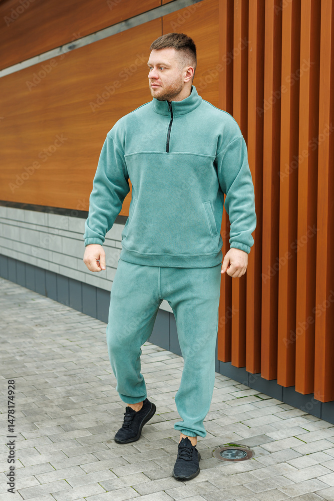 A stylish young man is confidently posing outdoors, dressed in a cozy teal fleece tracksuit. The backdrop features modern architecture set in an urban environment, creating a stunning visual appeal