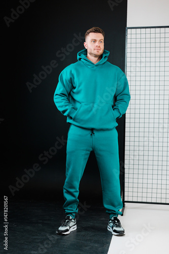Obraz na płótnie A fashionable male model is showcasing a vibrant teal hoodie paired with stylish joggers, all set against a modern and contemporary background that complements his activewear aesthetic perfectly w salonie