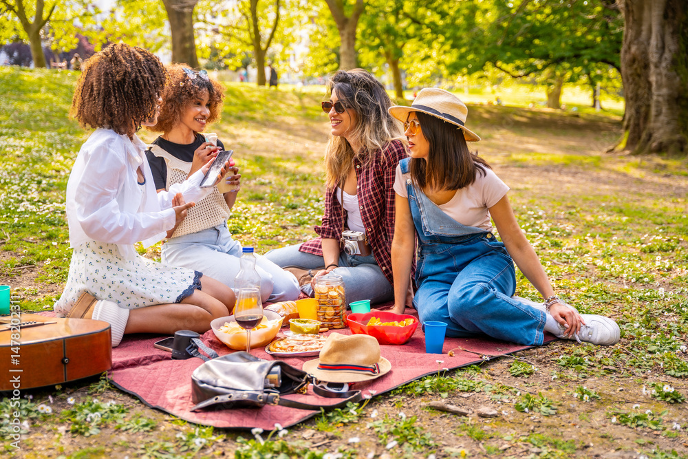 Cheerful women enjoying picnic, relaxing on blanket, using tablet and chatting in park Cheerful women enjoying picnic, relaxing on blanket, using tablet and chatting in park