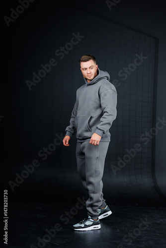Obraz na płótnie Casual Sportswear Lifestyle A Young Man in a Stylish Grey Hoodie and Comfortable Joggers Posed against a Dark Background, Representing a Trendy and Sporty Fashion Statement for Active Individuals w salonie