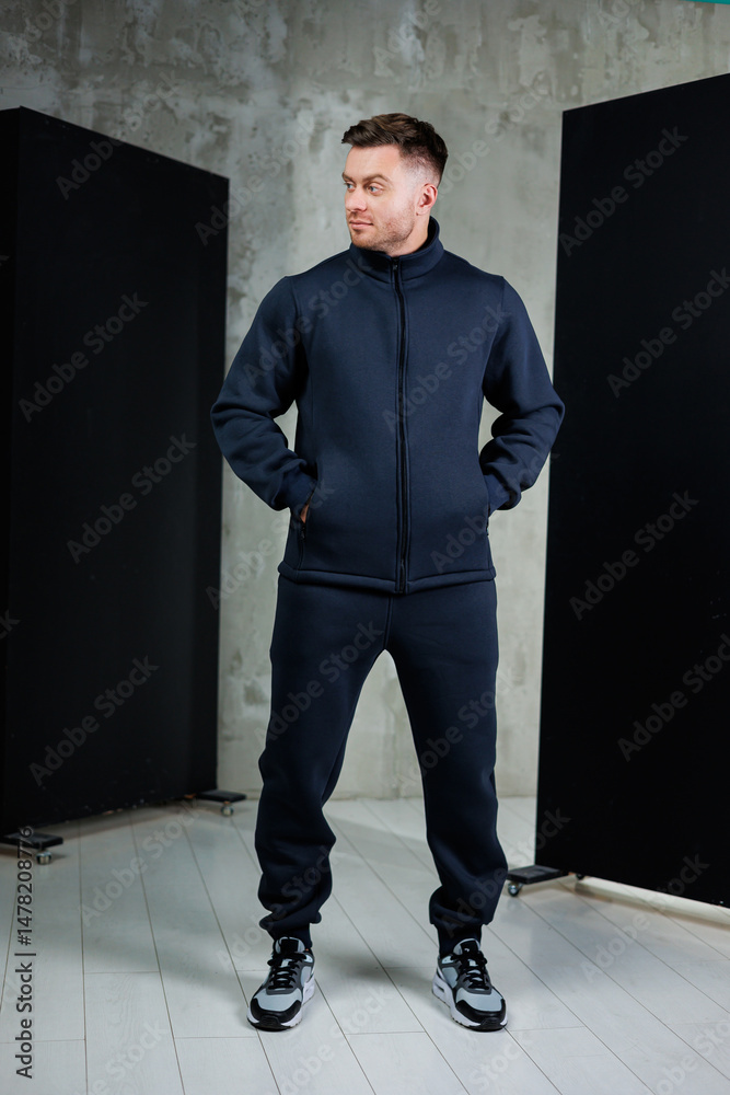 In a modern studio, a stylish man in dark sportswear exudes confidence while posing, showcasing a blend of aesthetics and current fashion trends with his activewear and trendy sneakers