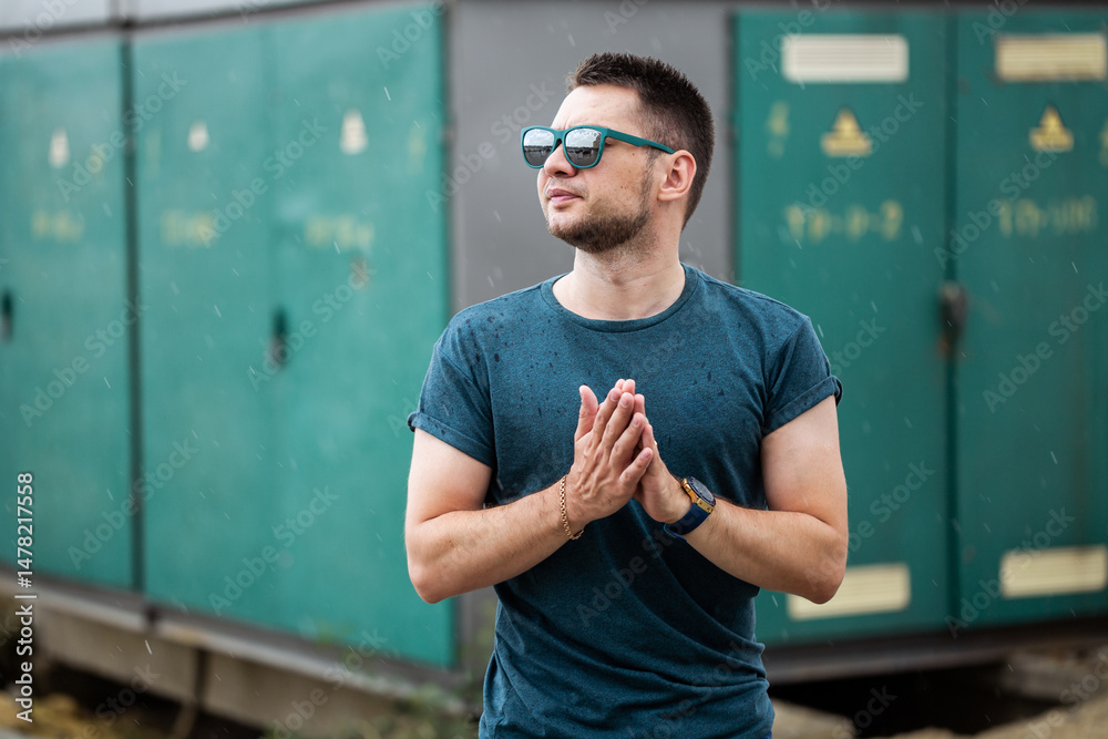 A young man in stylish sunglasses confidently poses outdoors in an industrial setting, surrounded by power units, exhibiting a relaxed, vibrant expression that reflects modern energy