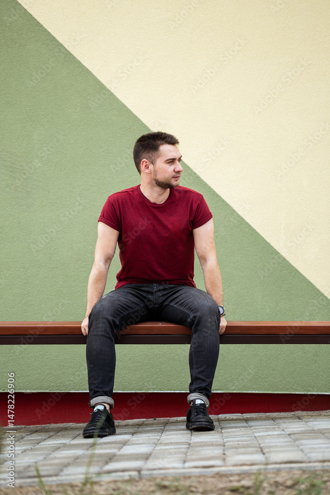 A young man dressed in casual attire is sitting comfortably on a bench that is positioned in front of a vibrant, colorful geometric wall, creating a striking contrast in the scene and ambiance