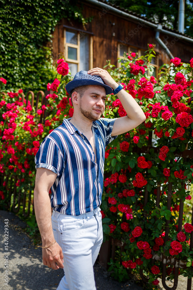 A fashionable man dressed in a stylish, striped shirt and wearing a trendy hat is enjoying a leisurely stroll amidst beautiful, vibrantly blooming roses during the delightful and warm summer season
