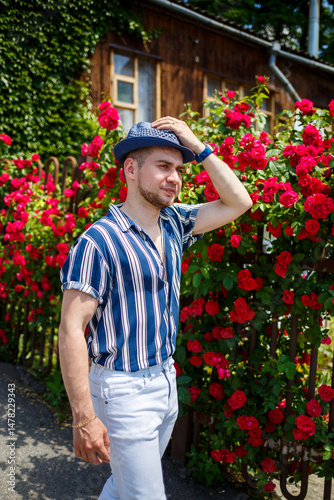 Obraz na płótnie A fashionable man dressed in a stylish, striped shirt and wearing a trendy hat is enjoying a leisurely stroll amidst beautiful, vibrantly blooming roses during the delightful and warm summer season w salonie