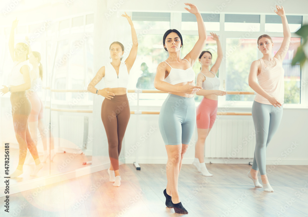 Naklejka premium Expressive young dark-haired woman in twenties, wearing workout clothes, mastering ballet moves at group class in sunlit dance studio