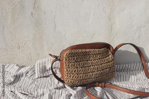 Woven straw and leather crossbody bag resting on striped fabric against white rustic wall background. Minimalist sunlit summer scene showcasing natural textures, stylish accessories. Copy space