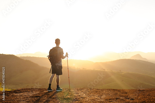 Young man hiking alone at sunset, standing on a mountain peak with trekking poles and a backpack, looking at the scenic valley below. Concept of freedom, solo travel and connection with nature