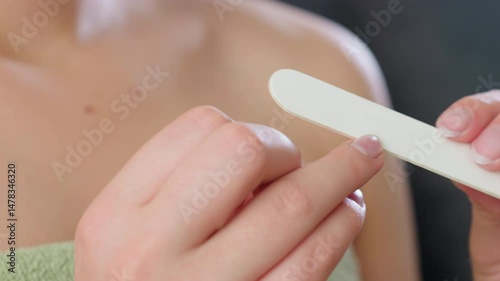 Close up hand view of woman gently filing fingernails with white nail filer while wearing green towel indoors, highlighting clean nail care, feminine grooming routine, and personal hygiene focus