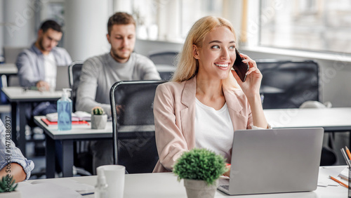 Modern work with client remotely in office with technology. Smiling businesswoman talking on smartphone and looking out the window, typing on laptop at workplace in office interior with workers, empty
