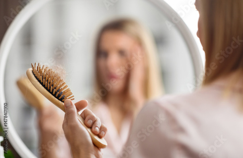 Hairloss Concept. Shocked Blonde Woman Holding Comb Full Of Fallen Hair After Brushing, Closeup Shot Of Middle Aged Lady With Brush In Hand Standing Near Mirror, Suffering Alopecia Or Health Problems