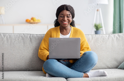 Photography Smiling young black woman in knitted sweater freelancer working from home, sitting on couch, using laptop, copy space