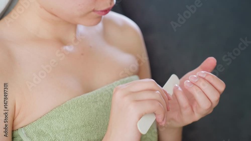 Close view of woman wearing green towel wrapped around chest, carefully filing nails with white nail filer in natural indoor light, capturing gentle selfcare moment with focus on hands and skin