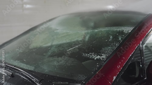 Rear view of person using high pressure car wash hose to rinse soap from car windshield inside garage with water droplets and suds streaming down glass surface during cleaning process