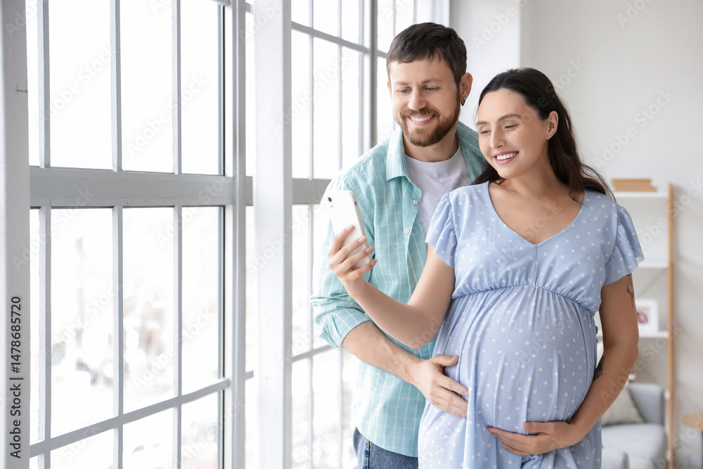 Happy pregnant couple using mobile phone near window at home Happy pregnant couple using mobile phone near window at home