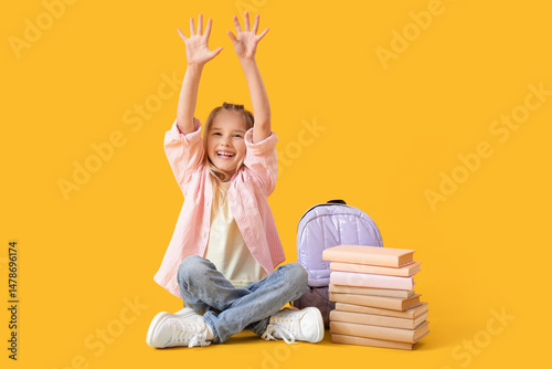 Little girl with books and backpack sitting on yellow background