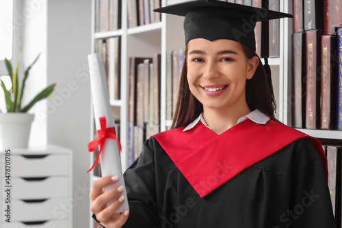 Smiling female graduate in cap and gown holding diploma in library, academic ...