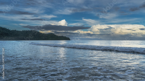 The ocean waves are calmly rolling towards the shore. Foam spreads across the beach. The clouds in the blue sky are illuminated with golden light. A hill in the distance. Early morning.Malaysia.Borneo