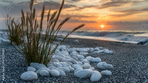 White pebbles and tall beach grass rest on a sandy shore as gentle waves roll in under a warm, colorful sunset sky, creating a serene coastal scene.