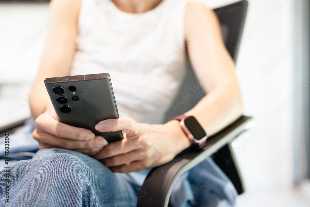 Naklejka premium Close up, low angle of woman’s hand holding smartphone, scrolling through social media, searching. She is in smart casual white sleeveless blouse, blue jean sitting in home office