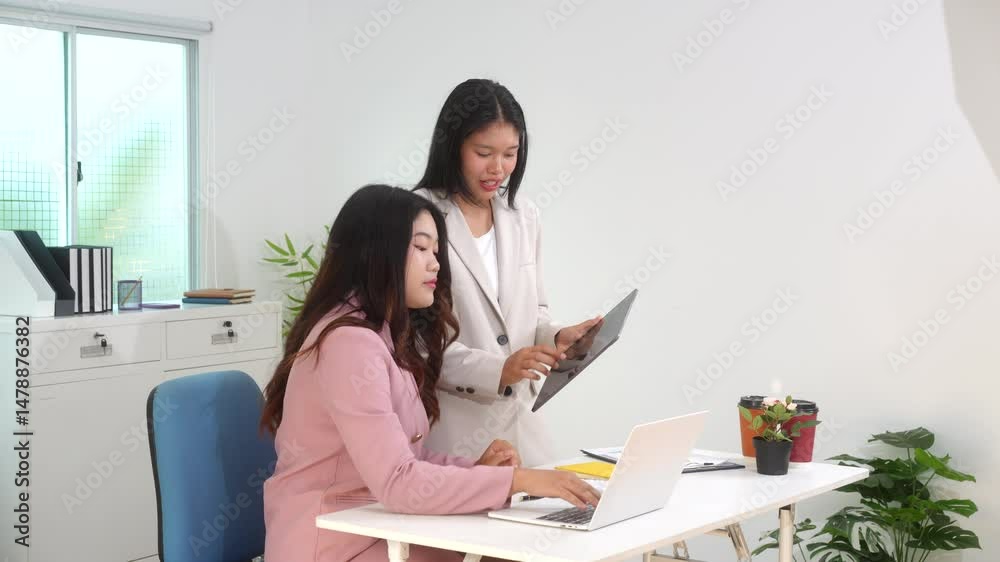 Two Asian business woman are discussing business reports and analyzing data at a desk. One is using a tablet while the other gestures. They appear focused,exchanging ideas in a modern office setting Two Asian business woman are discussing business reports and analyzing data at a desk. One is using a tablet while the other gestures. They appear focused,exchanging ideas in a modern office setting