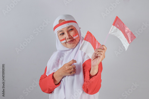 Joyful Muslim woman in hijab with face paint holding two Indonesian flags while smiling and pointing with pride during Independence Day celebration with nationalistic emotion and cultural pride
