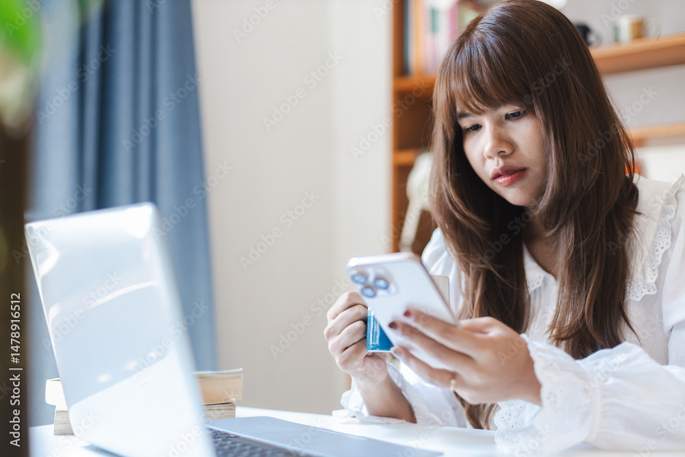 Asian female remote consultant is focused on her phone while working from home with laptop and books in cozy office space Asian female remote consultant is focused on her phone while working from home with laptop and books in cozy office space