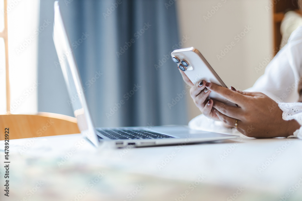Fototapeta premium Asian female remote consultant uses smartphone and laptop at bright workspace with natural light creating calm and focused atmosphere