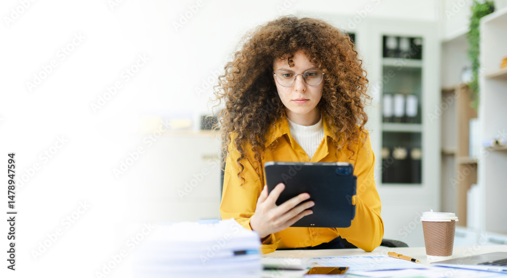 Obraz premium Confident businesswoman analyzing data on tablet at modern office desk with coffee, documents, and laptop. Concept of digital work