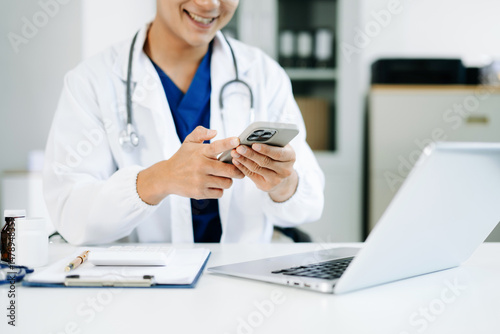 Schilderij op canvas Male doctor in uniform using smartphone at medical office desk with pills, clipboard, and stethoscope
