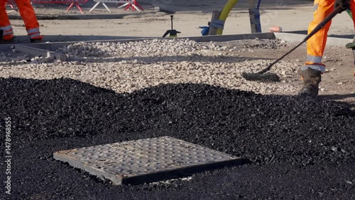 Road workers laying hot asphalt and prepare road surface around manhole cover at construction site