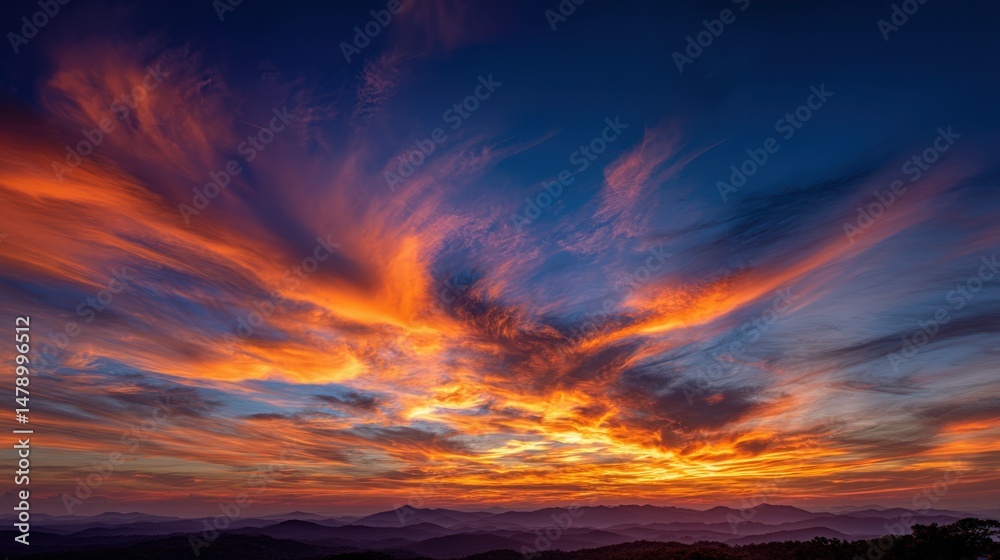 Naklejka premium Dramatic orange and blue sunrise lights up a layered mountain landscape with beautiful cloud formations in the sky