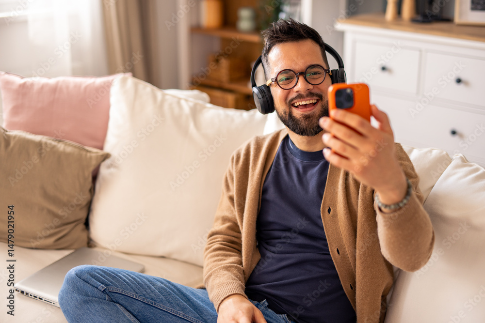 Obraz premium Smiling Man in Headphones Relaxing on Couch and Using Phone in Cozy Bright Living Room