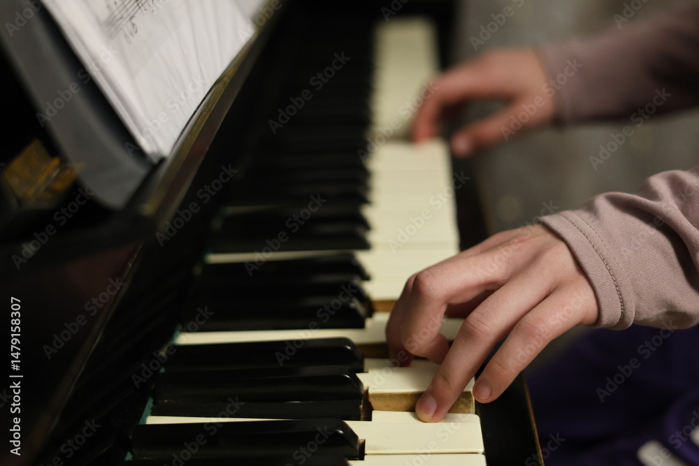Fototapeta premium Child or teen hands playing on a classic acoustic piano. Close-up of fingers on the keys, with sheet music visible in the background. The soft lighting and focus on hand movement create an atmosphere 