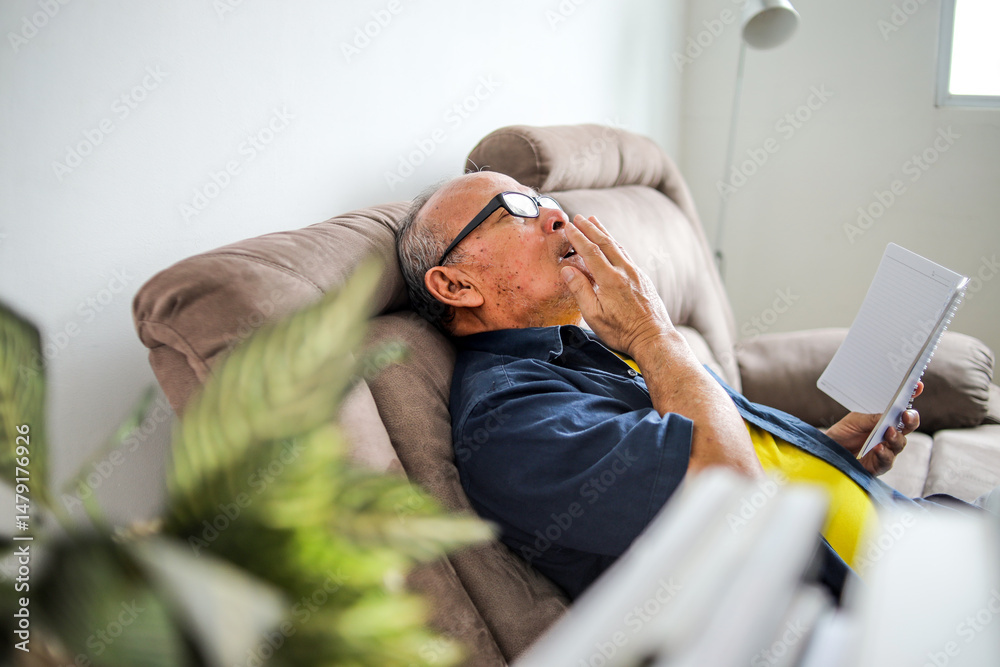 Fototapeta premium Sleepy Asian Aged Senior Man Yawning While Reading Book On Sofa