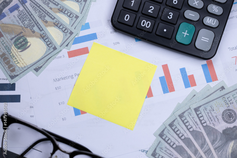 Fototapeta premium A yellow sticky note, cash, calculator, glasses, and financial charts on a desk, depicting business planning and financial analysis.