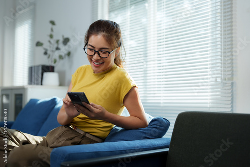 Happy asian freelancer woman using smartphone at home sitting on sofa