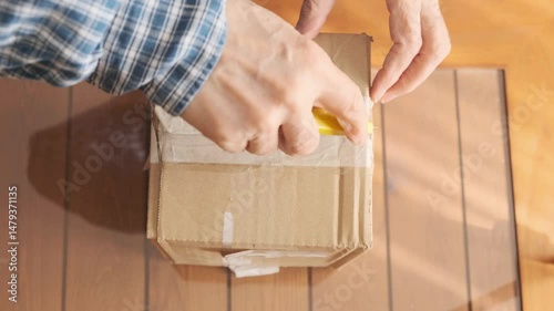 Opening a Sealed Cardboard Box Using a Yellow Utility Knife on Wooden Table.